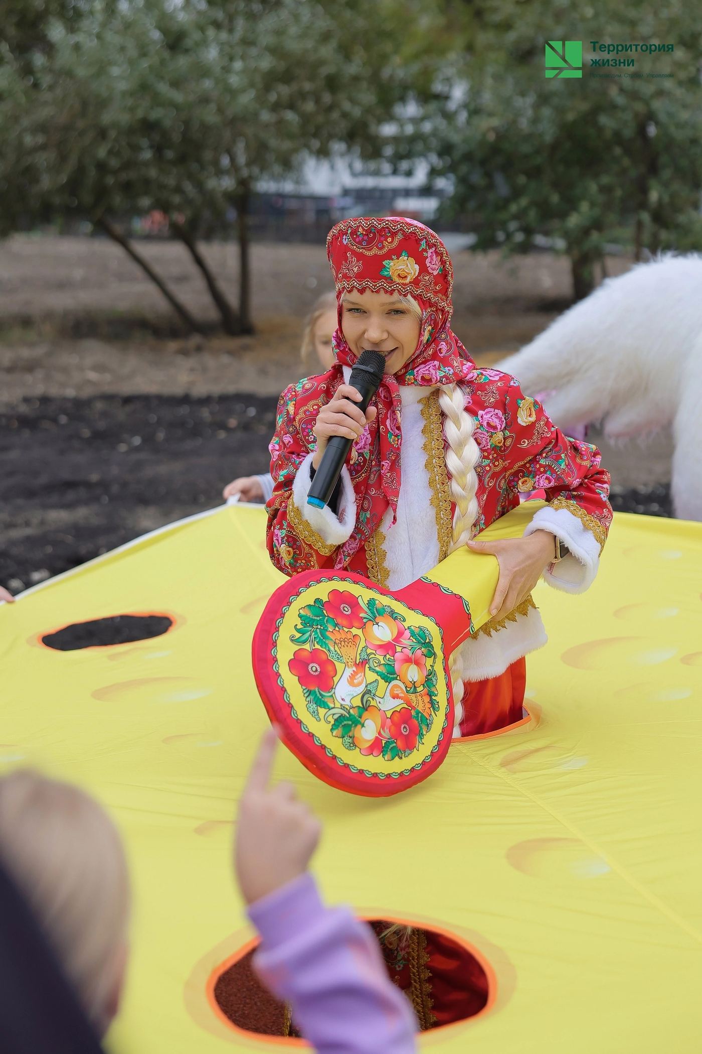 Уличная игра: ведущая в красном народном костюме с микрофоном и расписной балалайкой у жёлтого шатра